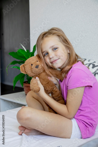 A cute little girl gently hugs a plush brown bear, looks at the camera and smiles while sitting on her bed at home.