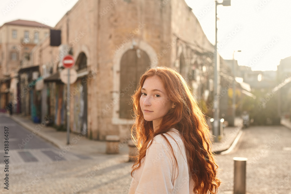 Fototapeta premium Young woman with red hair looking at the camera while walking in the city alone