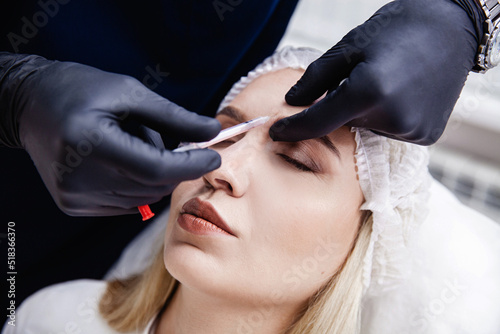A cosmetologist in black gloves injects botulinum toxin into the patient's forehead. A blonde patient in a disposable cap lies with her eyes closed