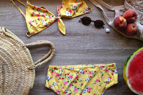 Yellow floral swimming suit, sunglasses, mesh tote with fresh peaches, seashell, watermelon half and wicker bag on wooden background. Summer essentials, flat lay.