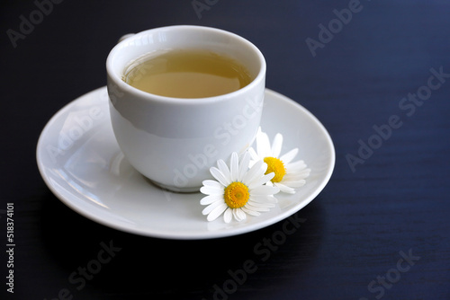 Chamomile tea in a white cup, daisy flowers on saucer. Healing herbal drink on dark wooden table
