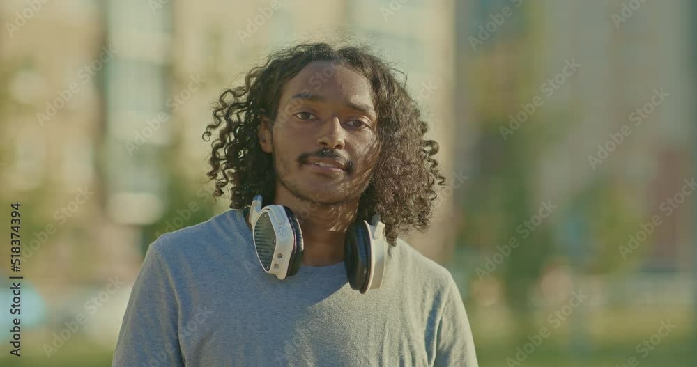 Portrait, dark-skinned handsome guy looks at the camera, he is happy. Guy with long hair and headphones smiles on a city street. 4k, ProRes Portrait, dark-skinned handsome guy looks at the camera, he is happy. Guy with long hair and headphones smiles on a city street. 4k, ProRes