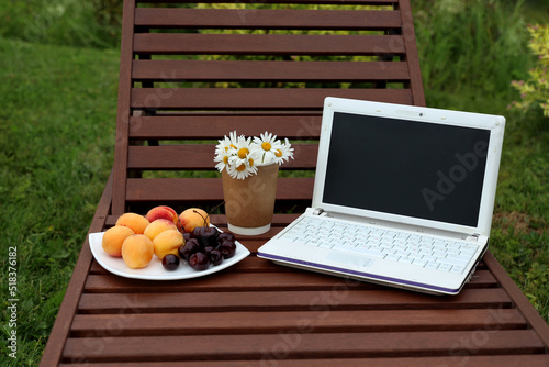 The blogger's attributes in summer are a white laptop, apricots and cherries on a plate, daisies in coffee cups standing on a wooden beach chaise longue