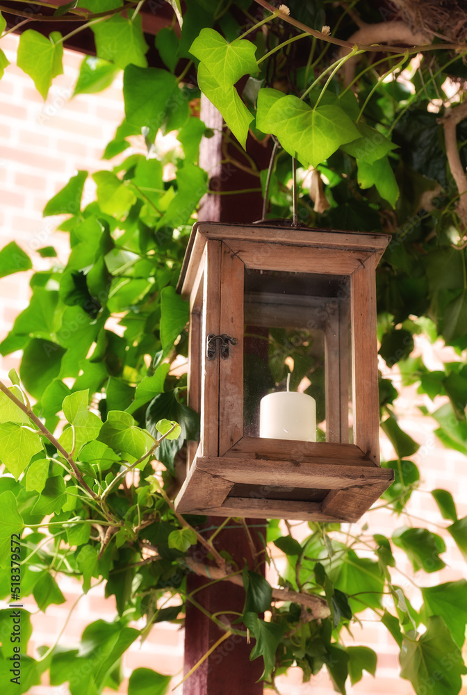 Burning candle in a lantern surrounded by green leaves in a garden or patio outside. Wooden frame hanging from roof for decoration and warmth in an outdoor entertainment area with lush plants