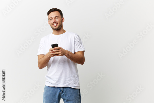 Cheerful tanned handsome man in basic t-shirt typing message with phone smile at camera posing isolated on over white studio background. Copy space Banner Mockup. Distance online communication concept
