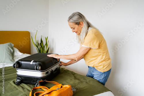 Gray-haired senior modern woman opening suitcase on bed photo