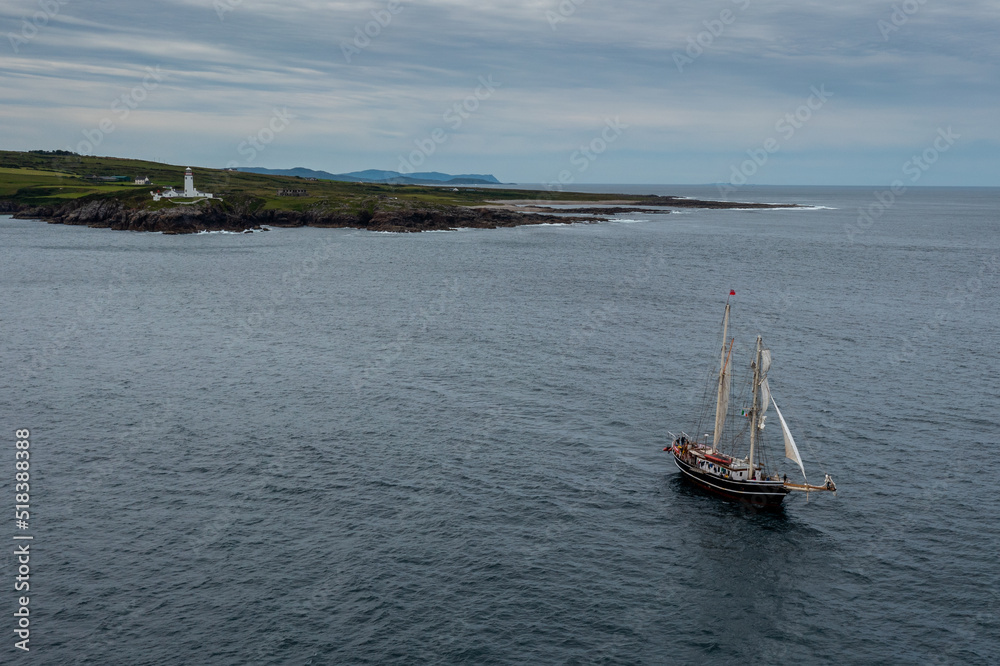 view of the Fanad Head Lighthouse on the northern coast of Ireland with an old two-masted sailboat sailing away view of the Fanad Head Lighthouse on the northern coast of Ireland with an old two-masted sailboat sailing away