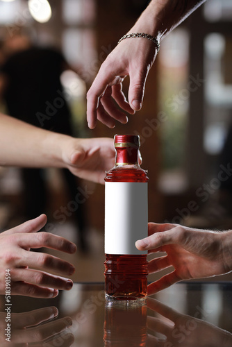 Glass bottle with alcohol drink with mockup on mirror table. Male people's hands reach for the bottle. Concept of celebration holiday in pub