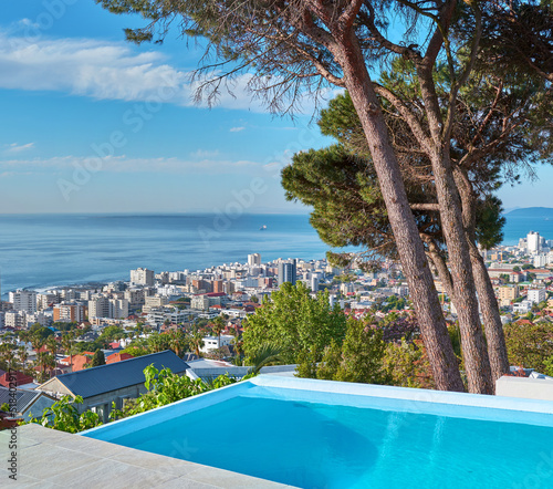 A blue pool on a luxury property with a coastal view of Cape Town and tourist landscape. The ocean and city against a clear sky horizon. A popular travel destination for tourists in South Africa