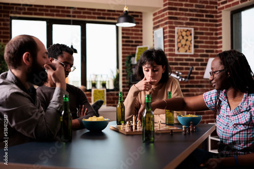 Smart multiethnic group of people sitting at table while playing chess. Happy diverse friends sitting together at home in living room, enjoying strategy boardgames while having beverages and snacks.