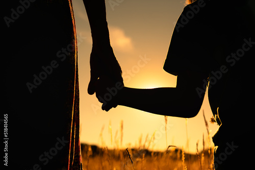 Mother little child silhouette holding hands walking in a grass field at sunset
