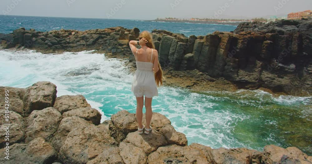Fuerteventura, Canary Islands. Slow motion. Relax happy smiling slim blonde woman, white dress, very long hair, standing on rocky ocean beach. Waves splash foaming spraying and hitting rocky cliffs. Fuerteventura, Canary Islands. Slow motion. Relax happy smiling slim blonde woman, white dress, very long hair, standing on rocky ocean beach. Waves splash foaming spraying and hitting rocky cliffs.