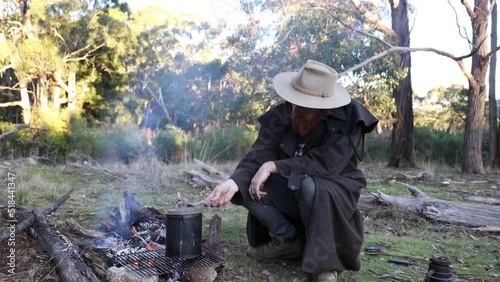 A bushman in an oil skin stockman jacket sits around a fire with his billy boiling in the high country. photo