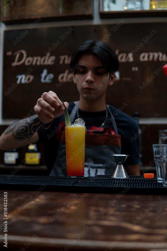 Bartender garnished a freshly brewed yellow drink with a green leaf. Waiter working at the bar with a drink for a client. Latino barman preparing a glass of juice