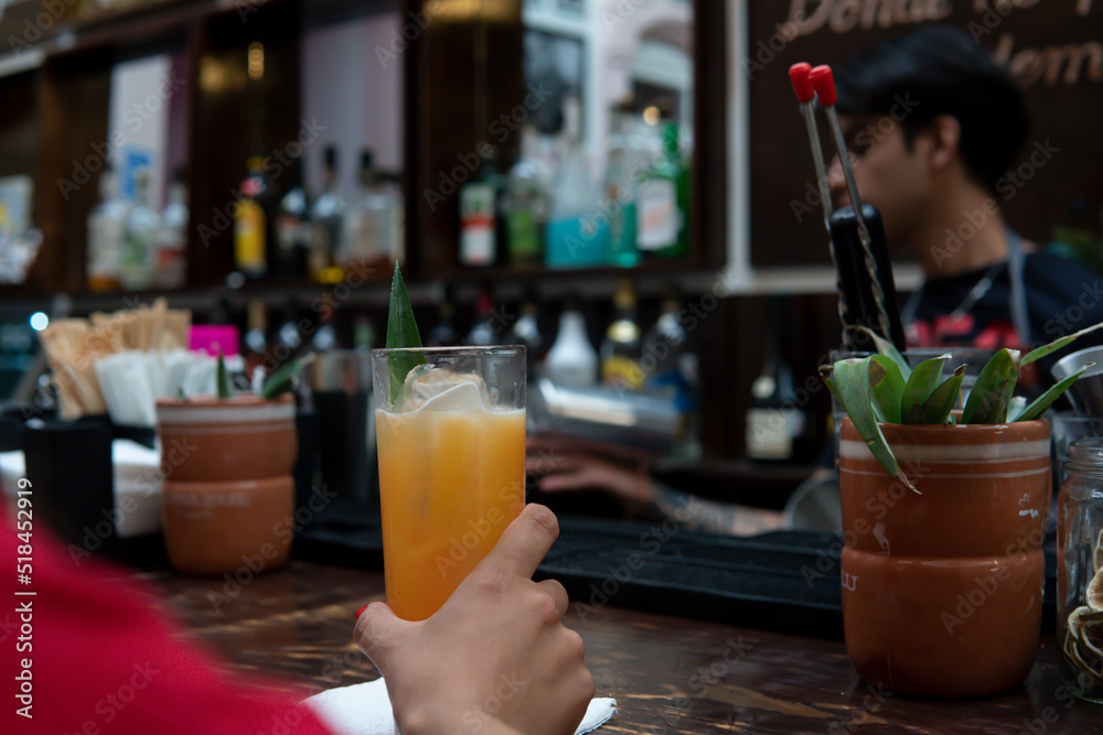 Hand of a girl in a red dress holding a glass of juice. Unrecognizable girl at a bar counter with the waiter in front of her. Wooden bar with rustic aesthetics of a restaurant.