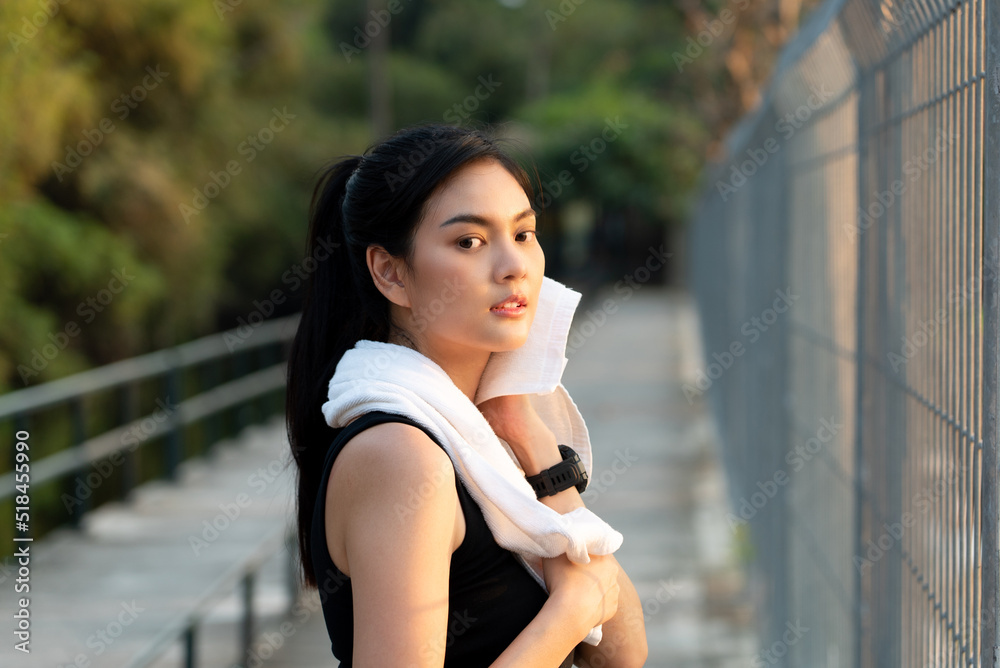 Close-up focus of a pretty Asian woman in sportswear standing on a walkway behind an Iron fence, wiping sweat on face with a towel on her neck while looking at camera after take a rest from running
