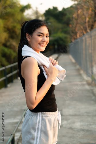 Vertical side view of young pretty Asian woman in sportswear standing on a walkway behind an iron fence, holding a towel draped around her neck smiling looking forward while take a rest after running.