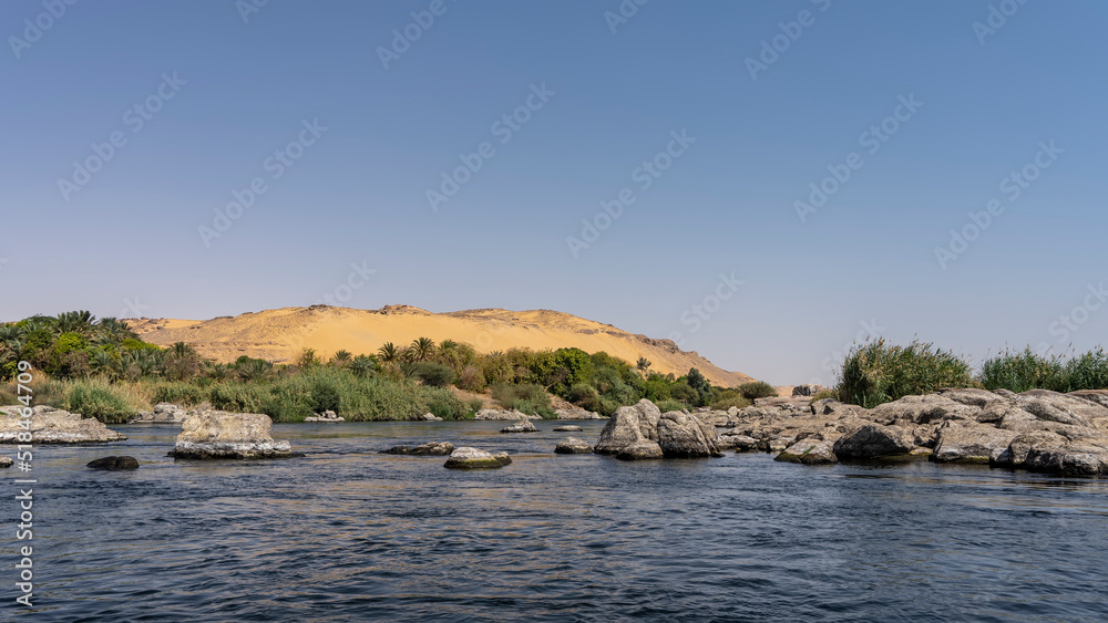 Picturesque boulders are visible in the riverbed and on the riverbank. Green vegetation on the banks. A sand dune against a clear sky. Ripples on the blue water. Egypt. Nile