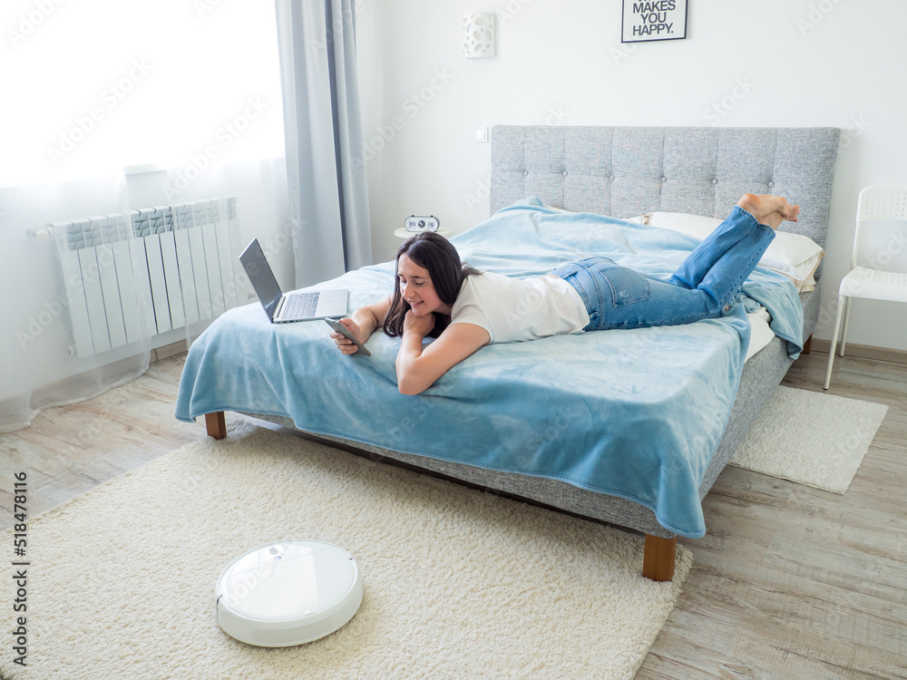 White robotic vacuum cleaner cleaning the floor while woman sitting on the bed and using phone. Smart technology concept. Woman in white t-shirt and blue jeans enjoying home cleaning. Work at home.