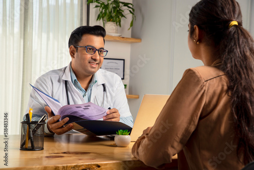Asian Indian male physician or doctor wearing white apron and stethoscope holding a file of medical report is consulting or prescribing medicines to a Young Female patient in modern clinic or hospital photo