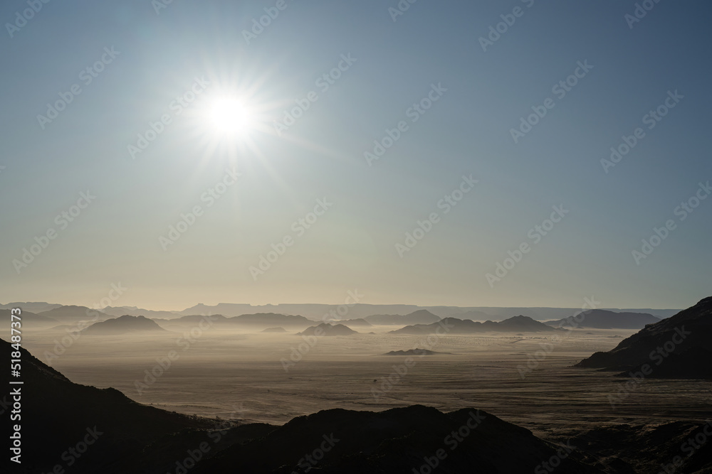 Silhouettes of hills and mountains in the Namib 
Desert in Namibia. The sun is high and clouds of windblown sand can be seen.