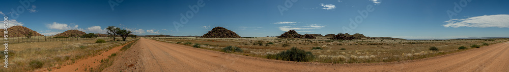 A panoramic photo of a gravel road in the middle 
of Namibia. Cairns with large boulders in the middle distance, blue sky with scattered white clouds.