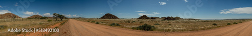 A panoramic photo of a gravel road in the middle of Namibia. Cairns with large boulders in the middle distance, blue sky with scattered white clouds.
