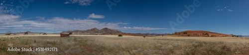 A panoramic photo of hills and mountains in central Namibia near Sossusvlei. First red dunes of the Namib Desert visible. Blue sky with scattered blue clouds.
