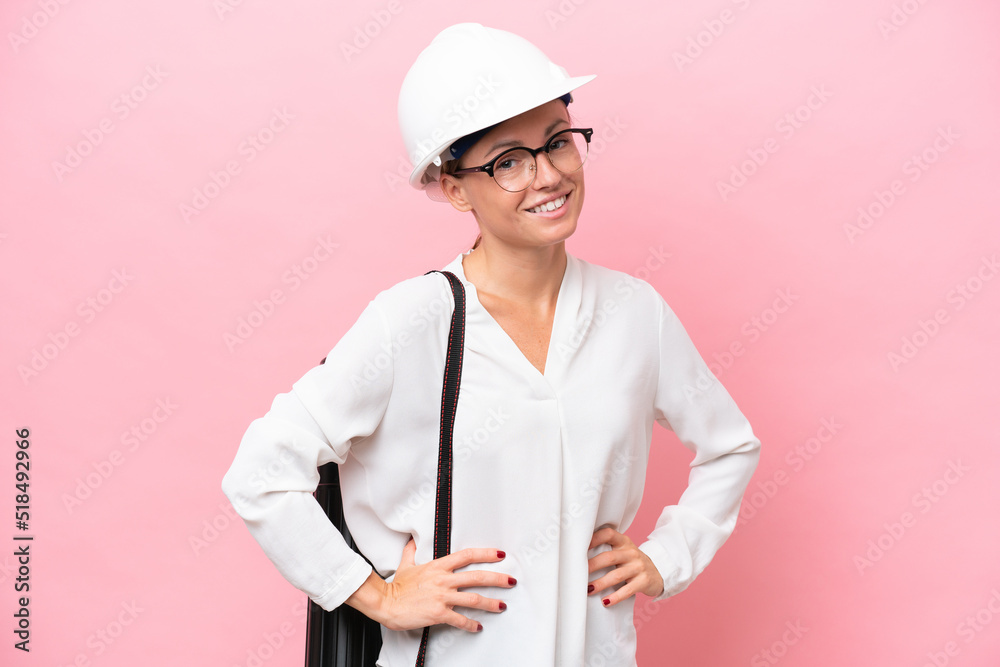 Young architect Russian woman with helmet and holding blueprints isolated on pink background posing with arms at hip and smiling Young architect Russian woman with helmet and holding blueprints isolated on pink background posing with arms at hip and smiling