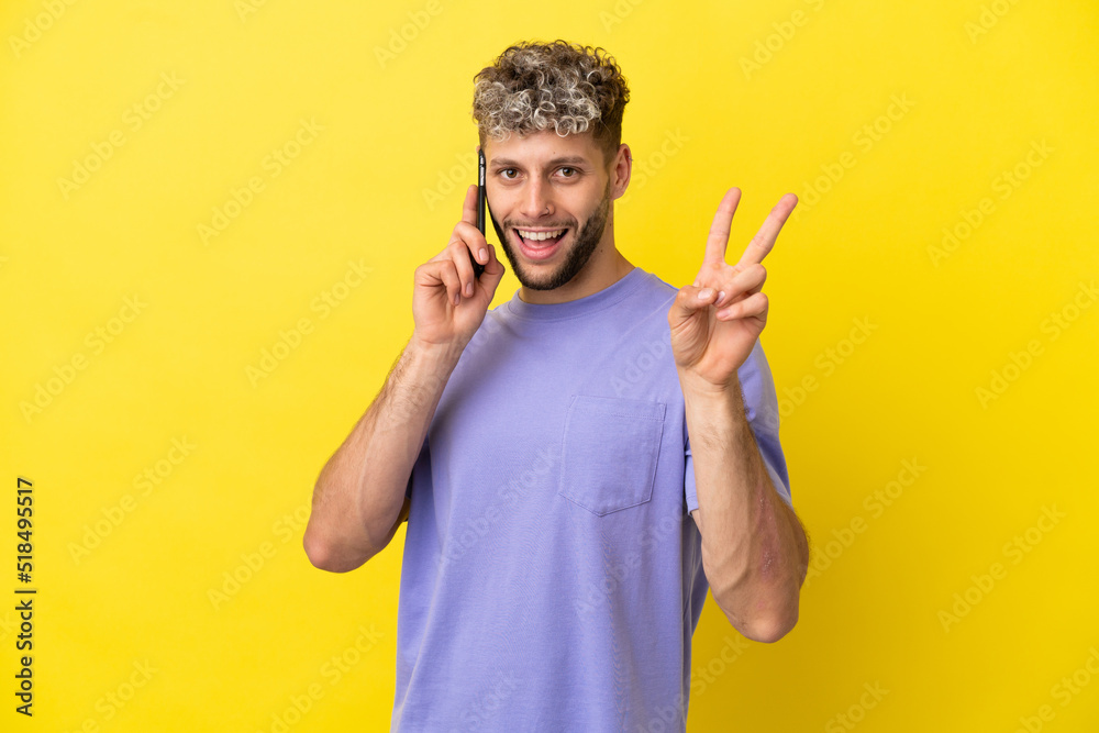 Young caucasian man using mobile phone isolated on yellow background smiling and showing victory sign Young caucasian man using mobile phone isolated on yellow background smiling and showing victory sign