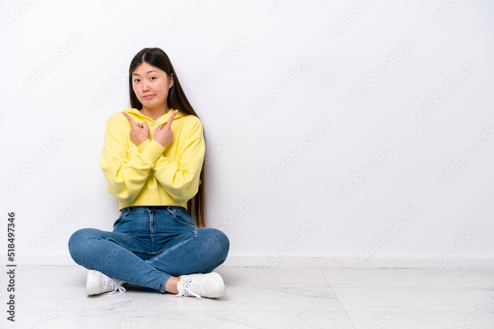 Young Chinese woman sitting on the floor isolated on white wall pointing to the laterals having doubts Young Chinese woman sitting on the floor isolated on white wall pointing to the laterals having doubts