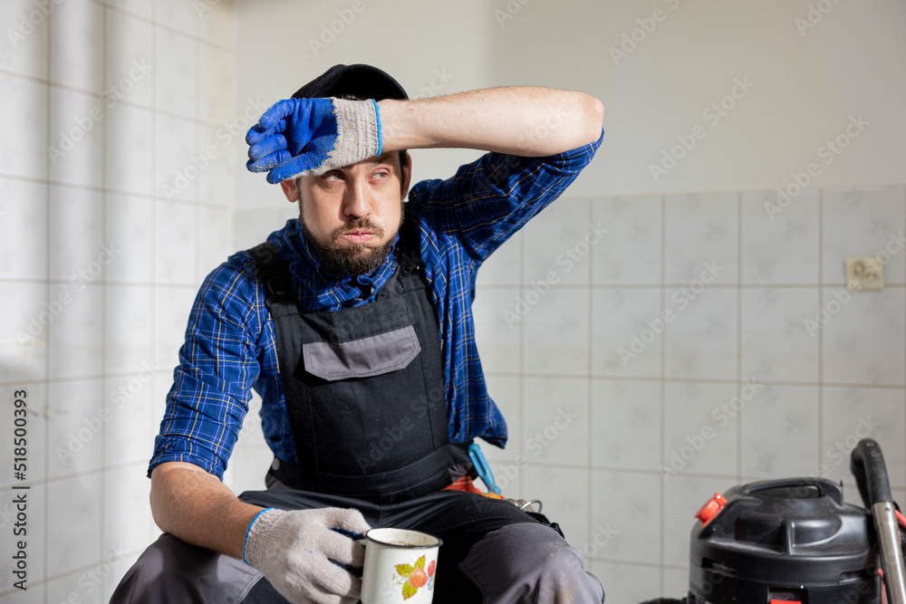A sweaty man tired of construction work sits in a renovated room on a stool wipes his forehead with a dirty glove with a sigh sips water from a mug, coffee.