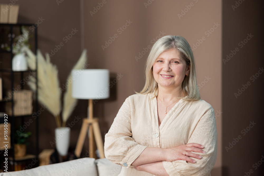 Headshot portrait positive mature woman with healthy smile standing on background of cozy living room, happy middle-aged lady looking to camera, posing for photo at home, enjoying leisure time. Headshot portrait positive mature woman with healthy smile standing on background of cozy living room, happy middle-aged lady looking to camera, posing for photo at home, enjoying leisure time.