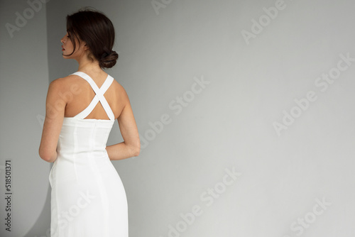 Back view of young woman brunette in white dress posing in studio, isolated grey background. Copy space.