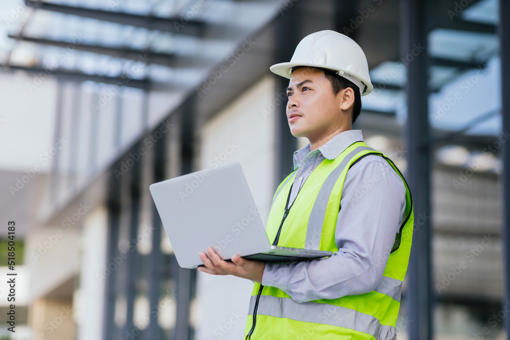 Asian engineer young man wearing safety vest and helmet standing using computer for check work on building construction site background. Engineering construction worker concept. Asian engineer young man wearing safety vest and helmet standing using computer for check work on building construction site background. Engineering construction worker concept.