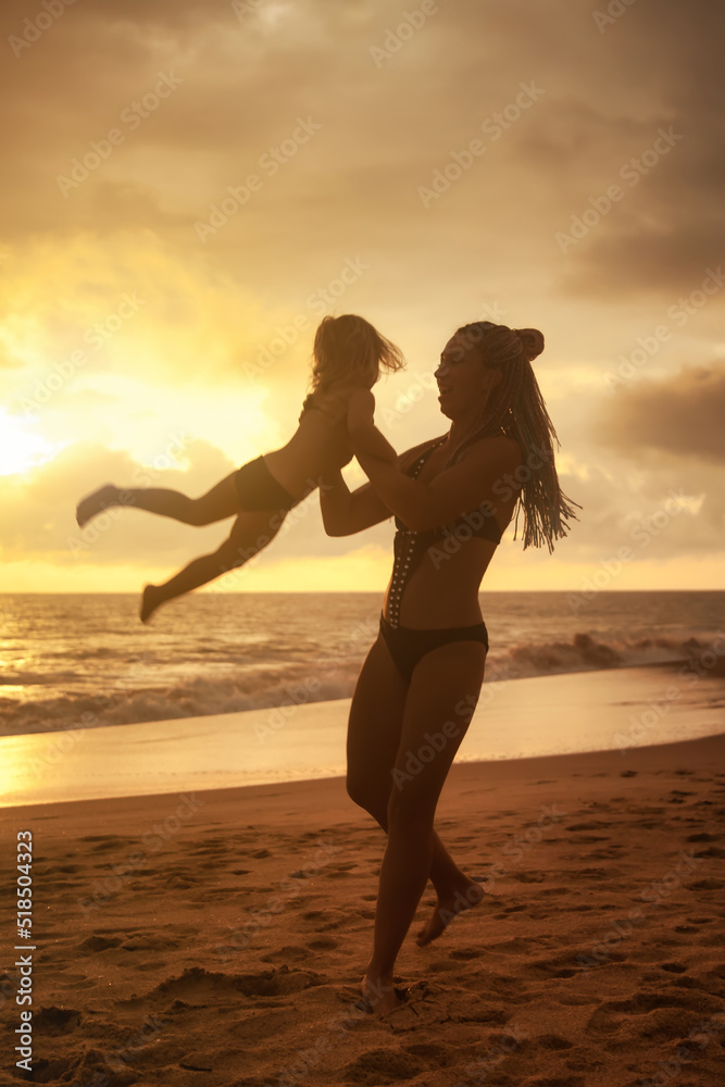 Silhouettes of mother and little girl daughter playing fun on beach at sunset sky background