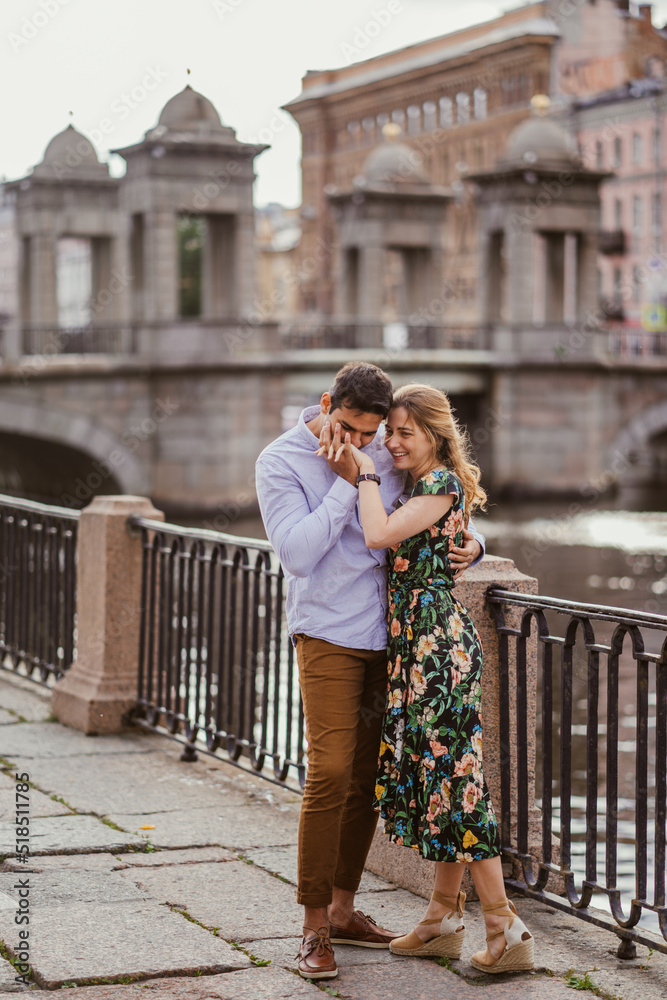 Brazilian young man kissing his blonde girlfriends hand in old town, against old bridge. Couple spending honeymoon, traveling. Lovers in casual clothes walking in the city. Relations and love concept.