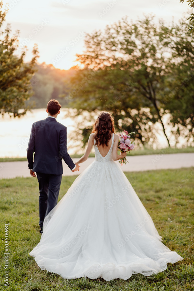 The bride and groom stand with their backs to us and look at the sunset against the background of trees and a river on their wedding day