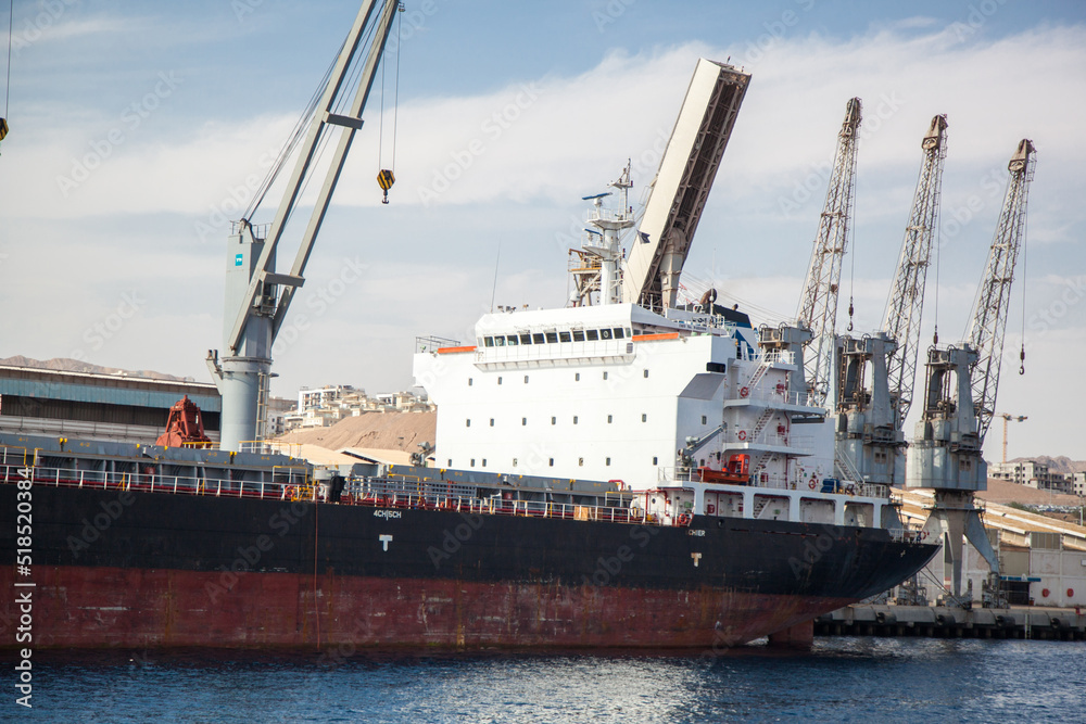 A large cargo ship is waiting for cargo to be loaded in the port. The large white cabin of the ship behind which the loading cranes of the port are visible. 