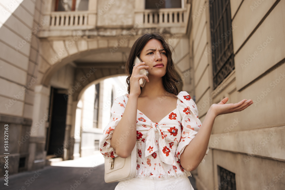 Fototapeta premium Outraged young caucasian woman talking on phone waving her hand standing on street. Brunette wears summer clothes on casual day. Telephone technology concept