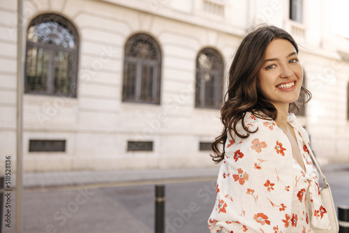 Smiling young caucasian woman posing for photo walking around european city during day. Brunette smiles with her teeth, wears white blouse. Summer vacation concept.
