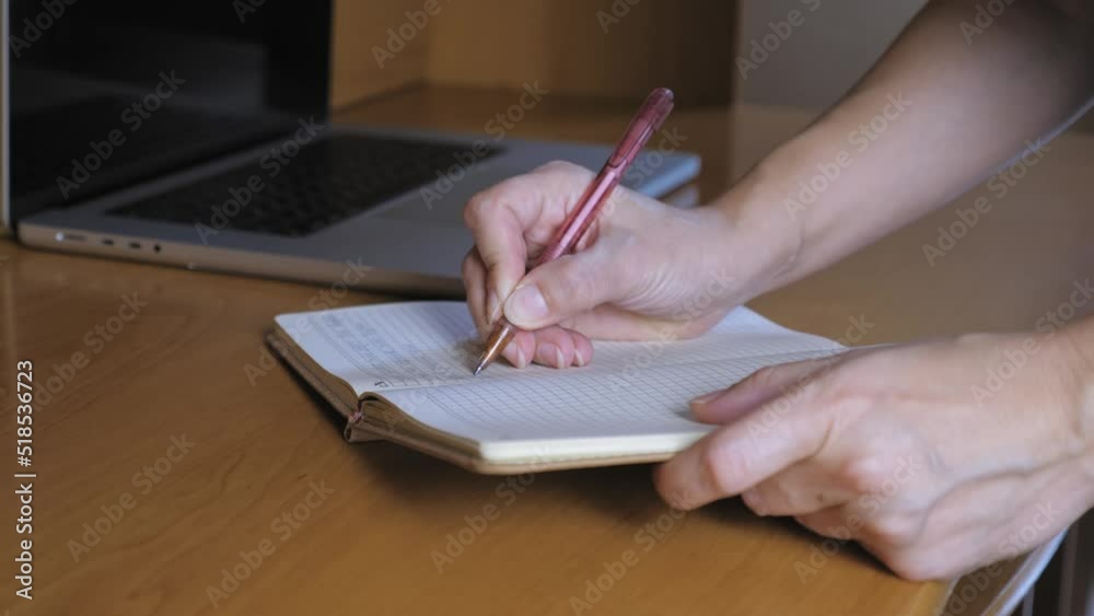 A woman writes in a notebook with a ballpoint pen. The notebook is on the table next to the laptop. Women's hands close up.