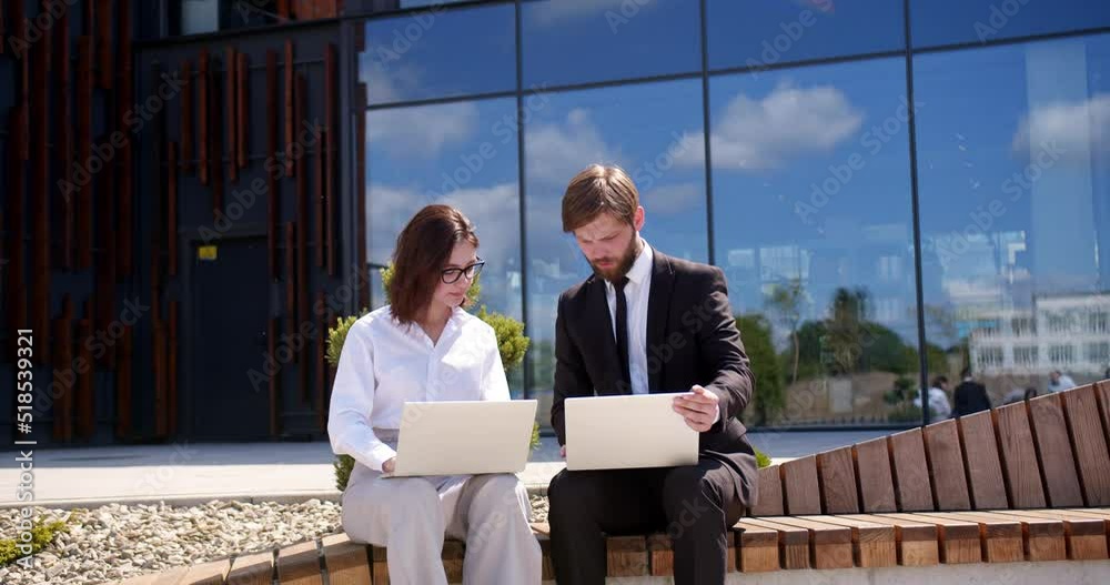Caucasian businessman and businesswoman sitting at street near modern office building and working on laptop computers while talking and discussing work. Man and woman having work discussion. Caucasian businessman and businesswoman sitting at street near modern office building and working on laptop computers while talking and discussing work. Man and woman having work discussion.