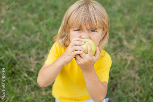 a happy blond boy is sitting on the lawn in a yellow T-shirt biting a green apple