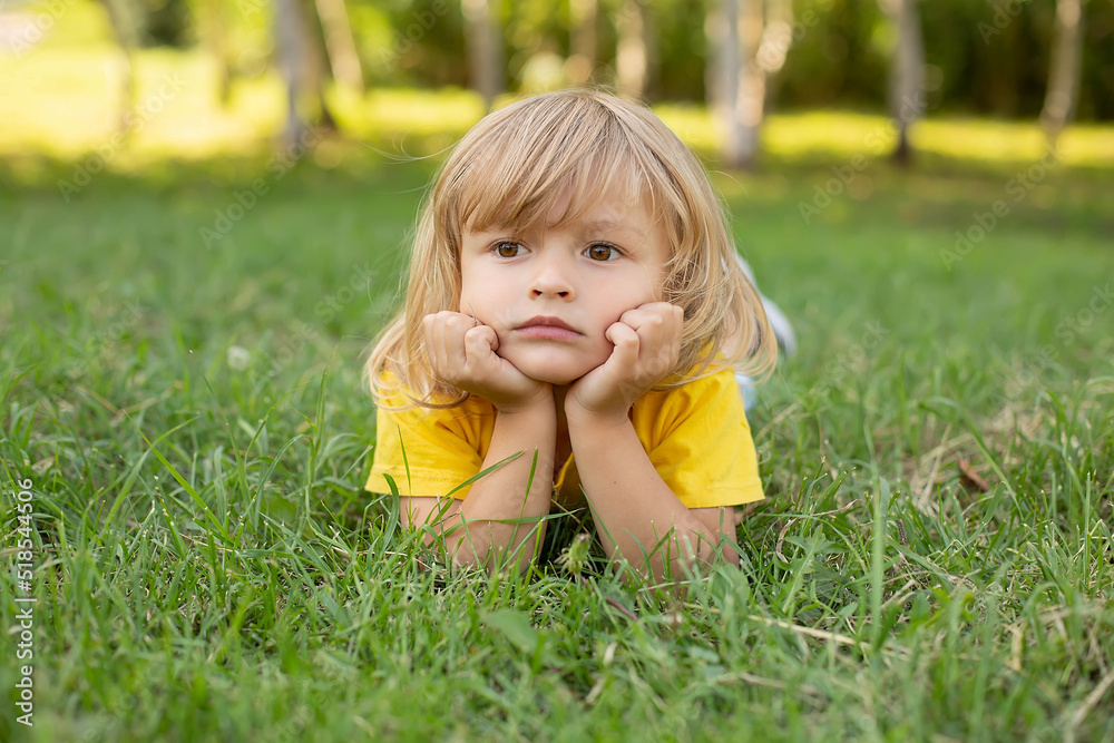 a blond boy with long hair and a mysterious thoughtful expression on his face lies on a green lawn a blond boy with long hair and a mysterious thoughtful expression on his face lies on a green lawn