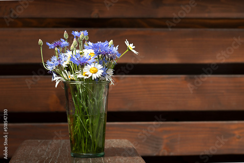 A bouquet of daisies and cornflowers in a glass vase on a wooden table against the background of a wooden wall. Place for an inscription. Selective focus. Copyspace.