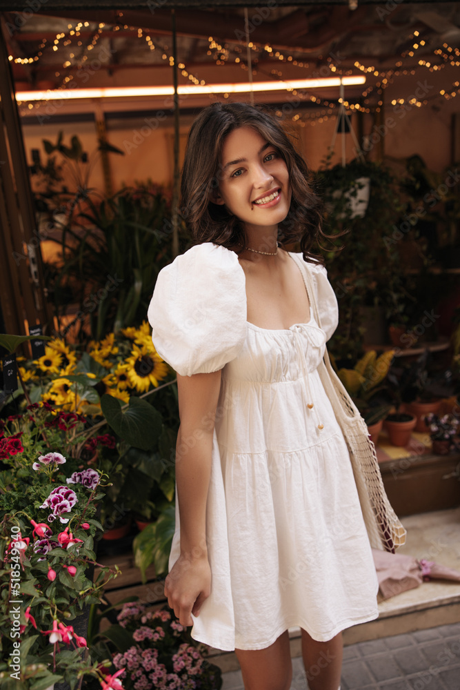 Cute young caucasian woman smiles with her teeth at camera standing on backdrop of flower market. Brunette woman with wavy hair wears white sundress in summer. Lifestyle, different emotions, concept.