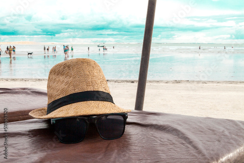 Fototapeta Naklejka Na Ścianę i Meble -  Hat and sunglasses on a beach chair with the sea in the background and blue sky. Brazilian beach. Summer holidays. Summer concept. Vacation travel concept. copy space