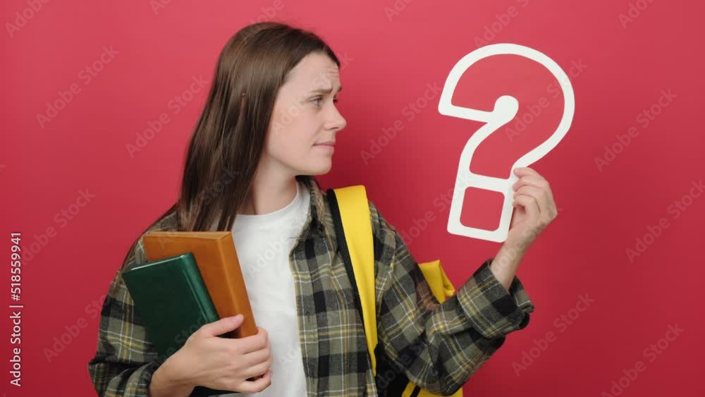 Puzzled pensive girl teen student holding question marks and books, wears shirt and yellow backpack, posing isolated over red color background wall in studio. Education in university college concept Puzzled pensive girl teen student holding question marks and books, wears shirt and yellow backpack, posing isolated over red color background wall in studio. Education in university college concept
