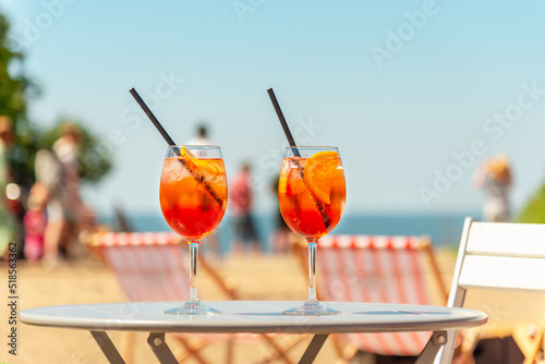 Two glasses of orange spritz aperol drink cocktail on table outdoors with sea and trees view blurred people background.